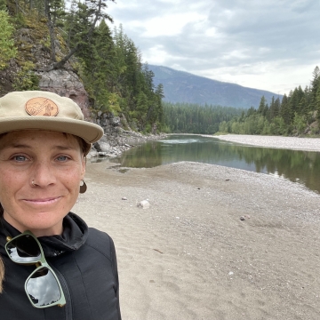 Picture of a women standing on a river shore with mountains in the background
