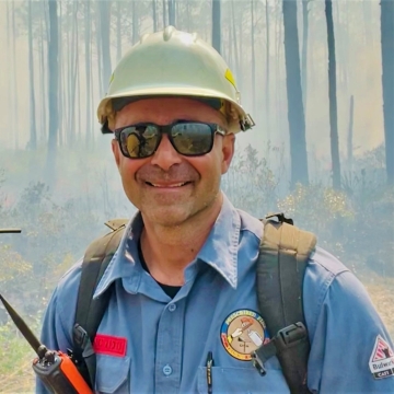 A picture of a man in a hardhat standing in the woods with smoke.